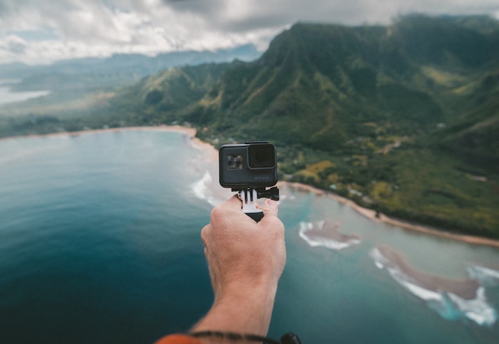 Person holding a GoPro brand camera in their hand pointed at a water and mountain landscape