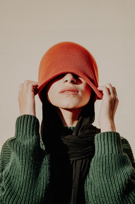 a portrait of a woman in a green sweater pulling an orange beanie over her eyes